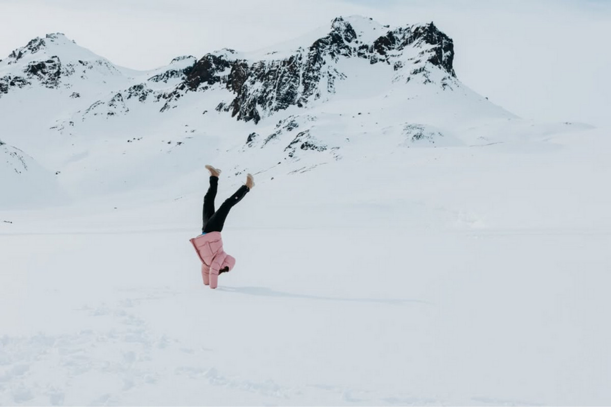 A person in a pink jacket doing a handstand on a vast, snow-covered landscape with large, rocky, snow-capped mountains in the background in Iceland.