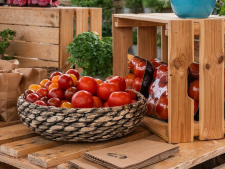 A display of ripe red and orange tomatoes in a woven basket and wooden crates at Sólskinsbúðin Farmers Market, with brown paper bags for customers.