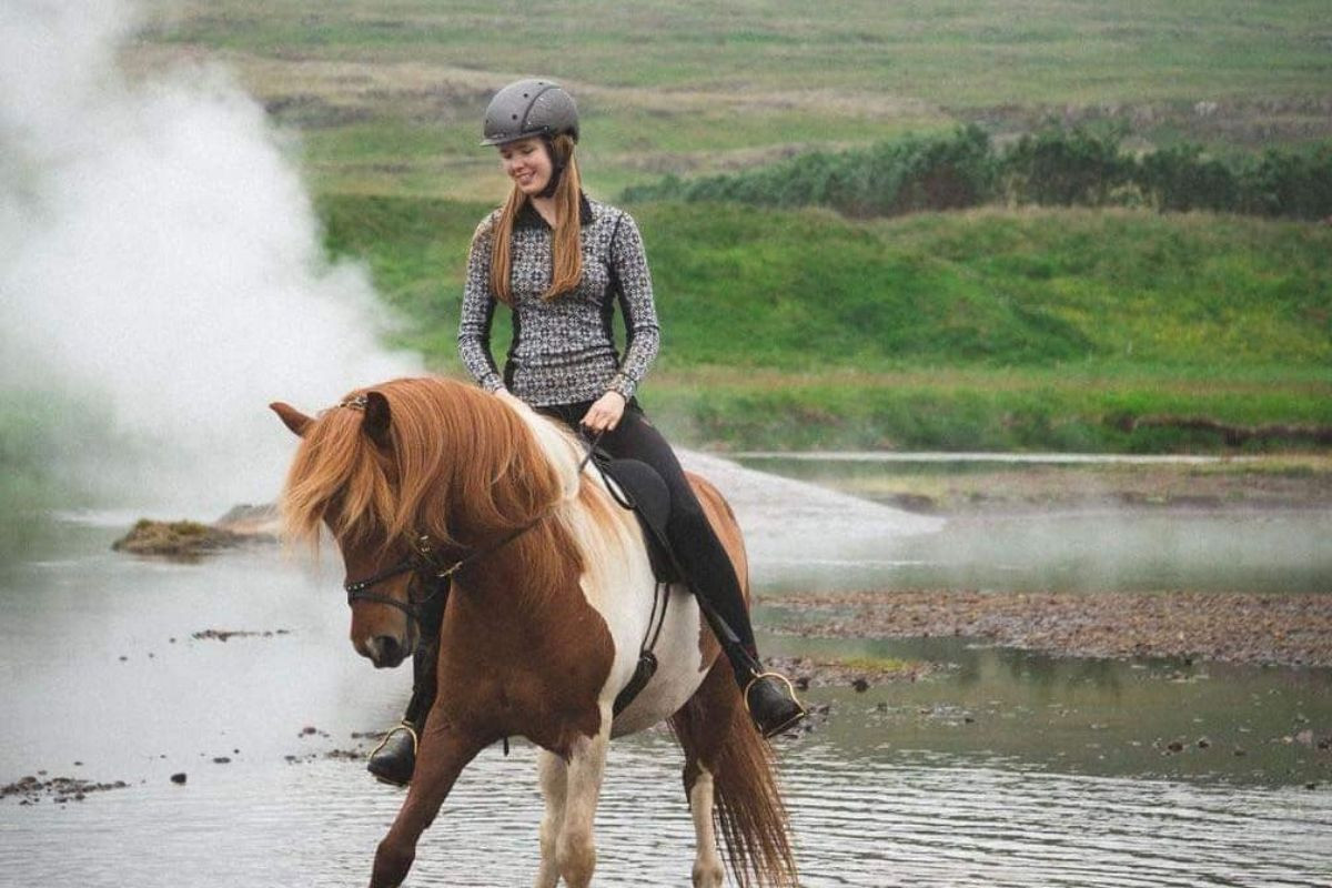 A woman smiling while riding a pinto Icelandic horse through shallow water with steam rising from a hot spring in the background at Sturlureykir Horse Farm.