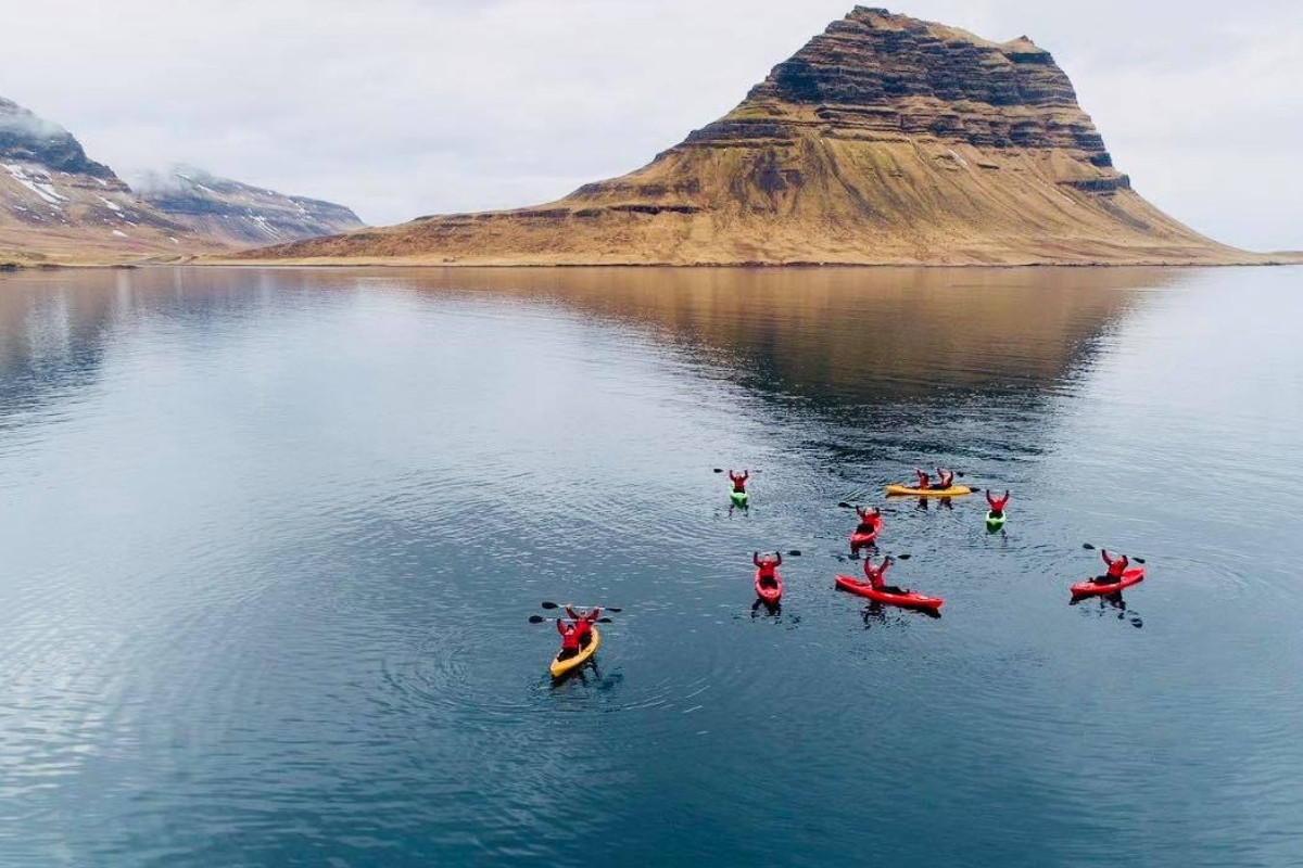 An aerial view of multiple red and yellow kayaks with people raising their paddles in a bay in Iceland, with the iconic Kirkjufell mountain in the background.