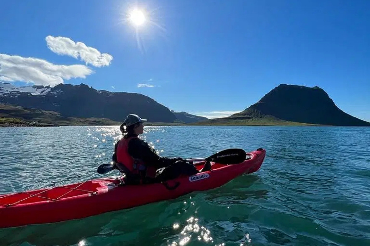A person in a red kayak on sparkling blue water, looking towards majestic mountains and glaciers under a bright sun in Iceland.