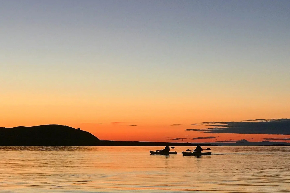 Two kayakers silhouetted against a vibrant orange and blue sunset sky on calm water in Iceland.