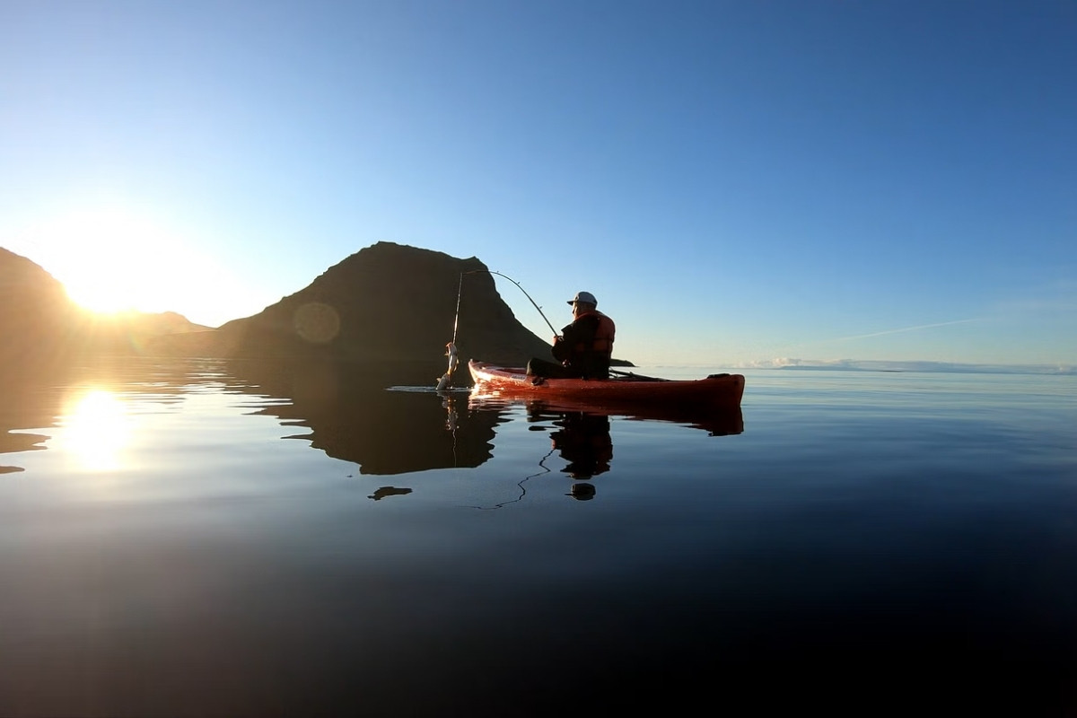 A lone kayaker fishing on calm water during sunrise in Iceland, with a dramatic mountain silhouette reflecting on the water.