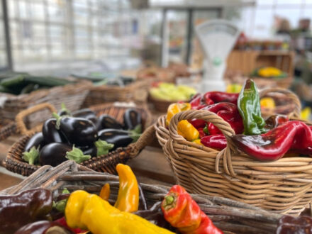 A close-up view of baskets and piles of fresh, colorful vegetables including red, yellow, and green peppers, and dark purple eggplants at a farmer's market.