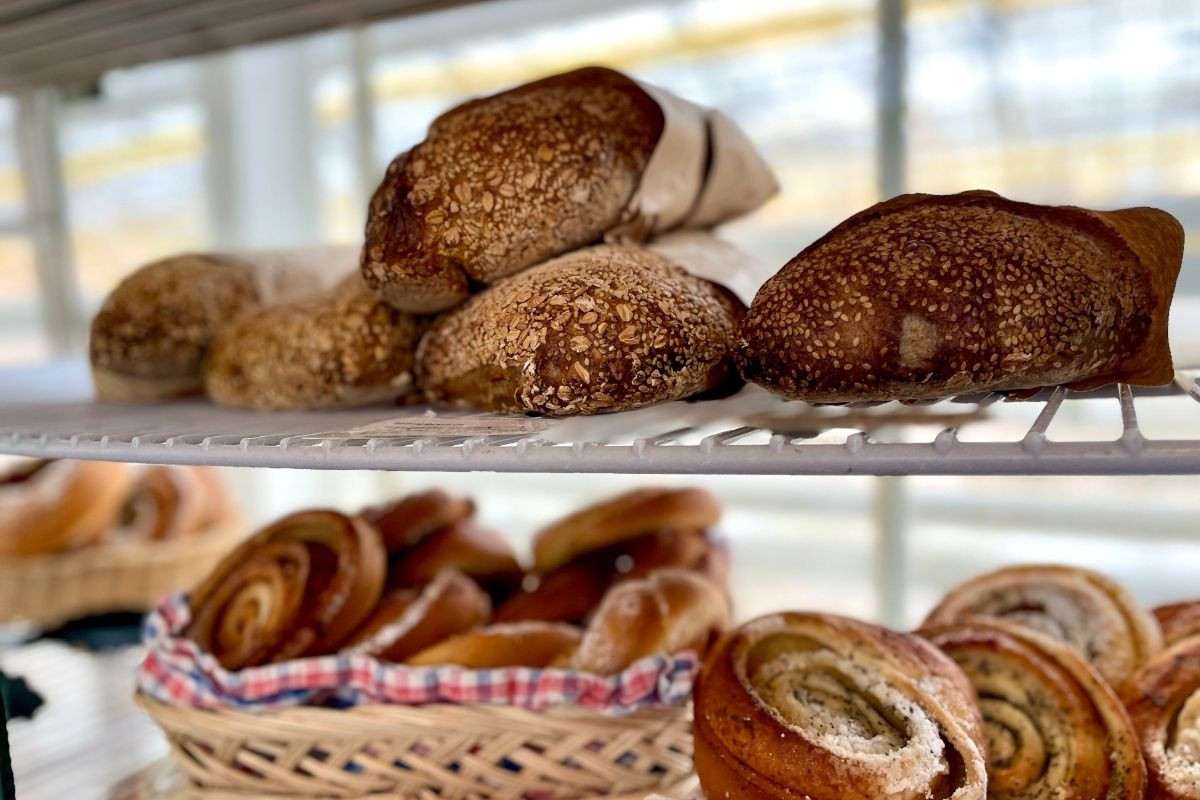 A bakery shelf holds various fresh baked goods, including loaves of dark, rustic bread wrapped in paper on the top shelf and several cinnamon rolls in a basket and on the lower shelf