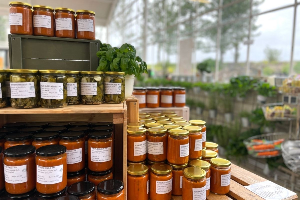 Jars of preserves and sauces are stacked on wooden crates and shelves, with a green plant in a pot on top of a crate in the middle of the display.