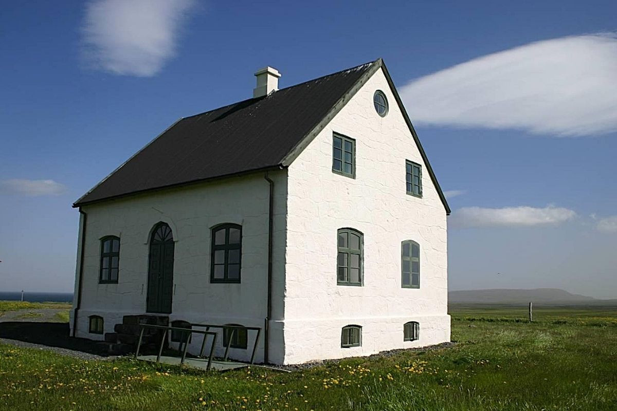 Sauðaneshús, a historic white stone farmhouse with a dark roof and green window frames, standing in a vast green field with a blue sky and distant ocean in Iceland.