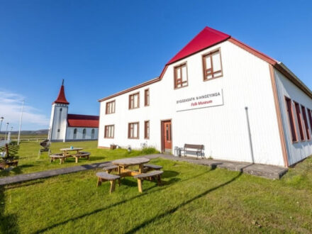 A bright white building with a red roof, labeled "BYGGÐASAFN N-ÞINGEYINGA Folk Museum," stands in a green grassy area with picnic tables. In the background, a white church with a matching red steeple is visible under a clear blue sky.