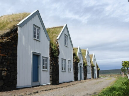 Row of traditional Icelandic turf houses with white wooden gables, turf roofs, and stone bases, under a cloudy sky.