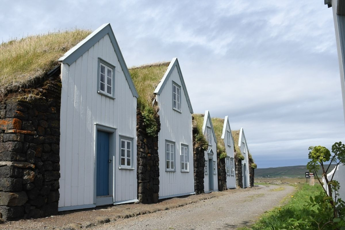 Row of traditional Icelandic turf houses with white wooden gables, turf roofs, and stone bases, under a cloudy sky.